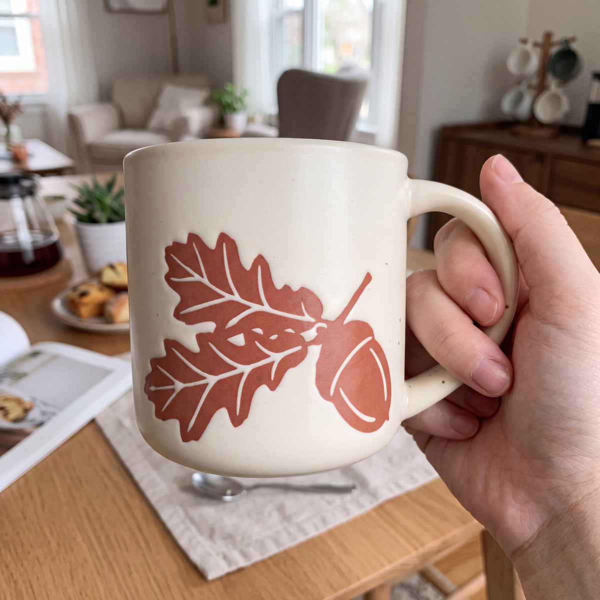 Hand holding The Acorn stoneware mug over an oak dining table during a cozy morning coffee routine