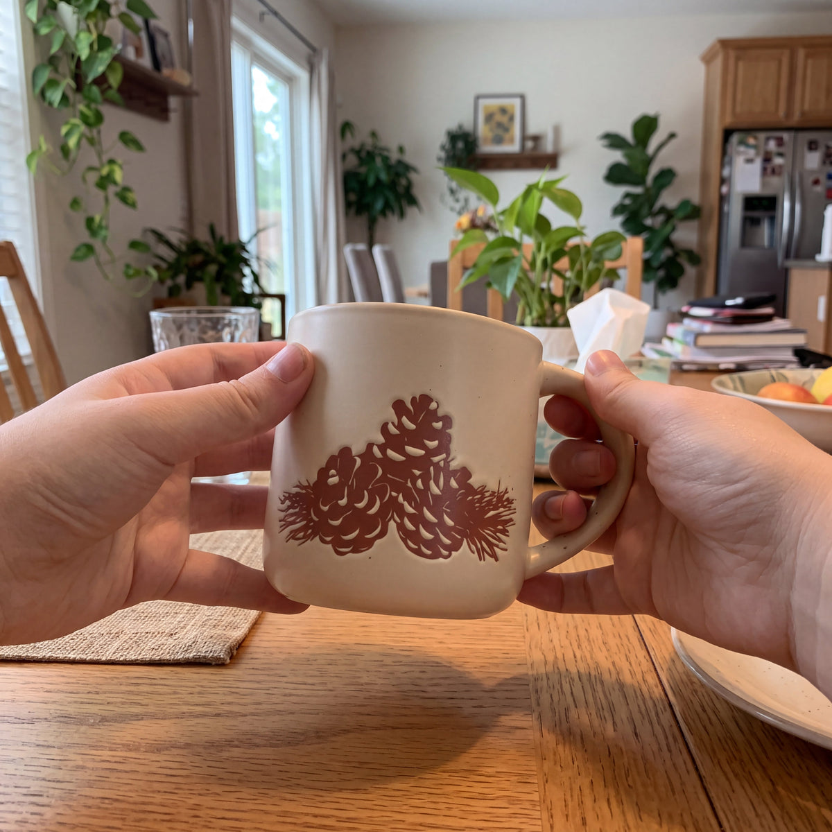 Two hands cradling The Pinecone ceramic mug over a warm honey oak table surrounded by lush houseplants