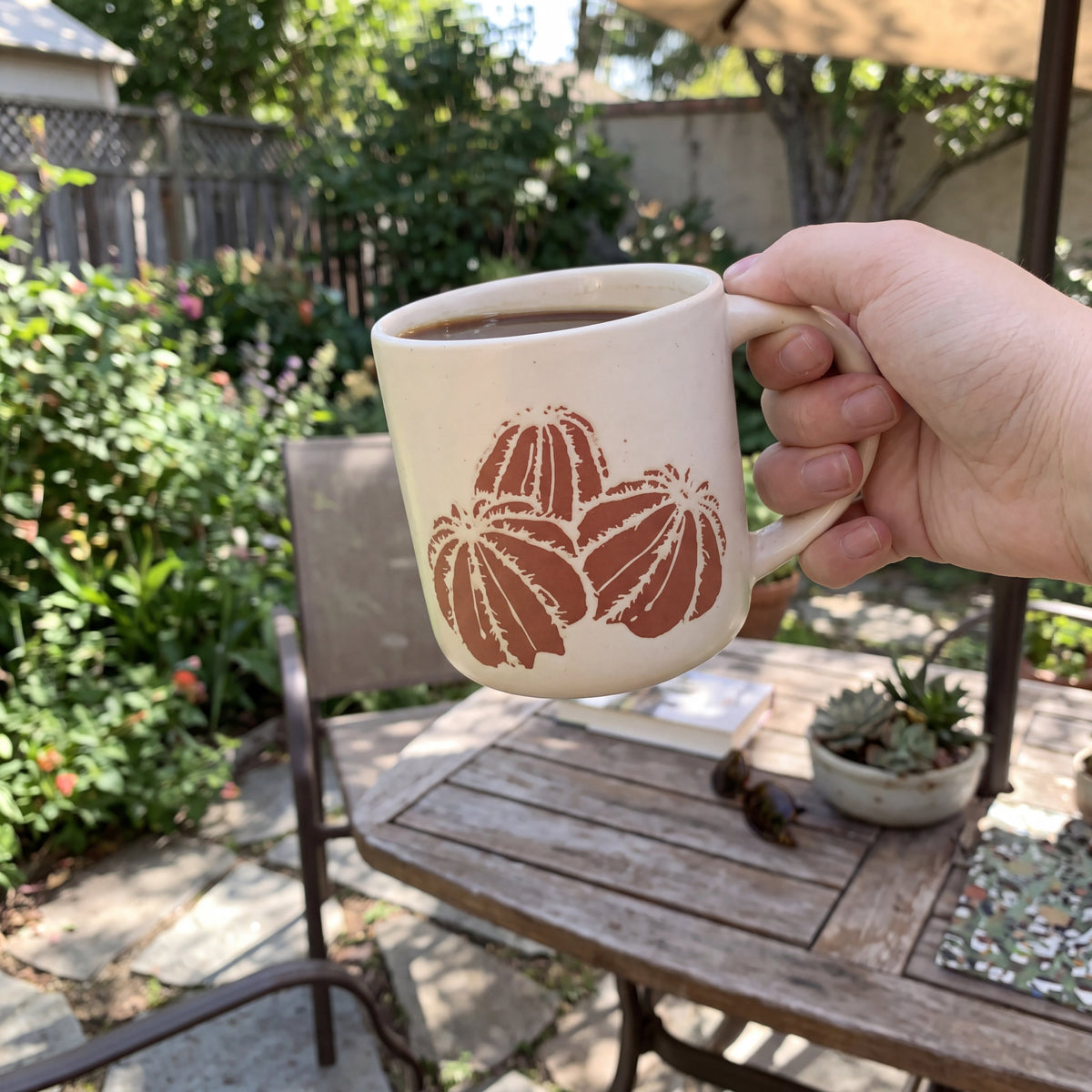 Hand holding The Cactus stoneware mug on a sunlit wooden patio table surrounded by outdoor greenery