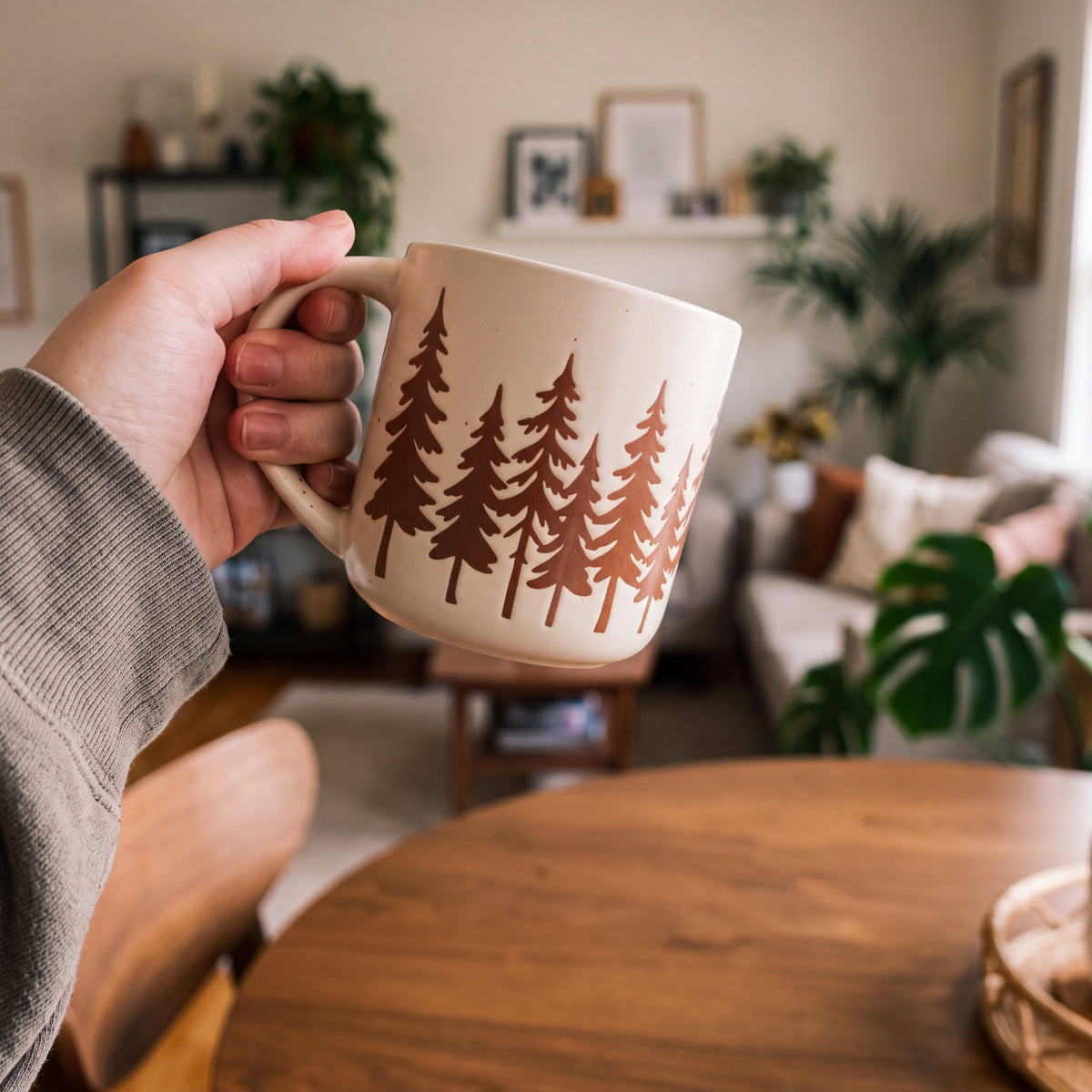 Hand holding The Evergreen ceramic mug over a round oak table in a cozy, plant-filled living room