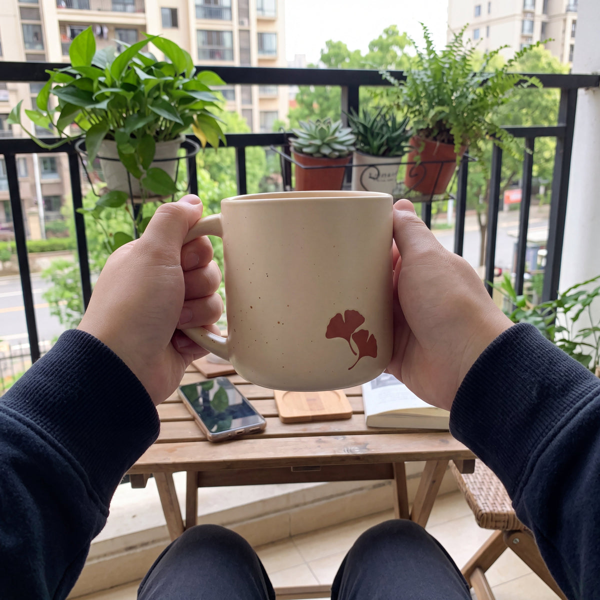 Two hands cradling The Ginkgo ceramic mug on a balcony surrounded by lush green potted plants