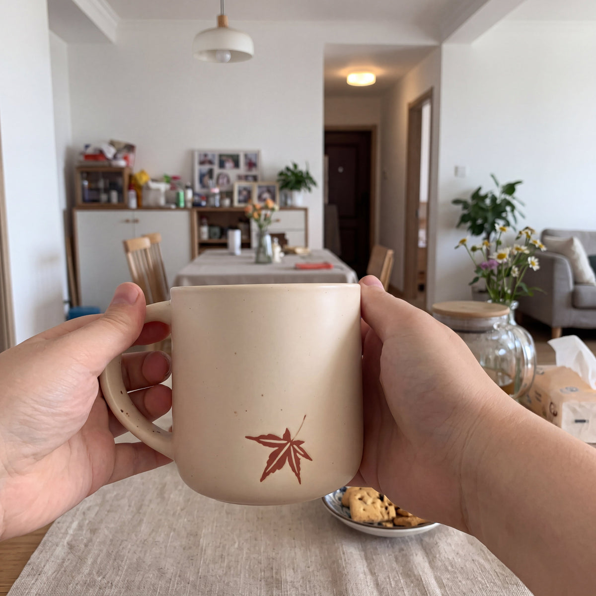 Two hands cradling The Maple ceramic mug over a warm linen dining table with baked cookies