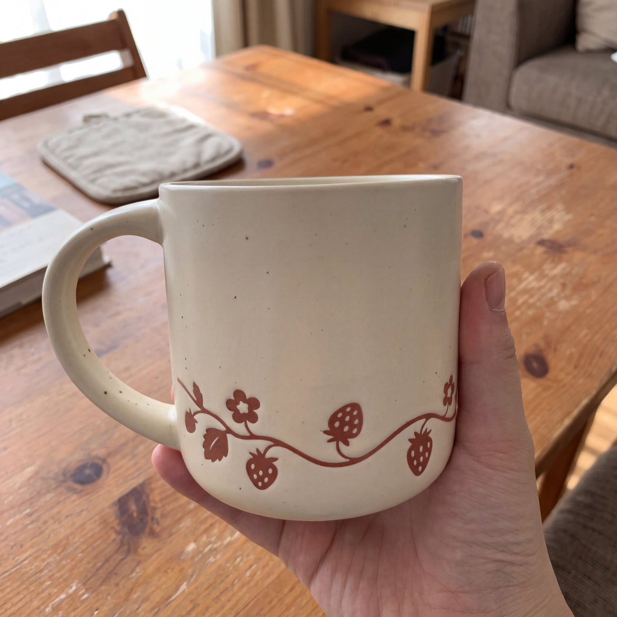 Hand holding The Wild Strawberry ceramic mug over a honey pine table during a slow morning coffee routine