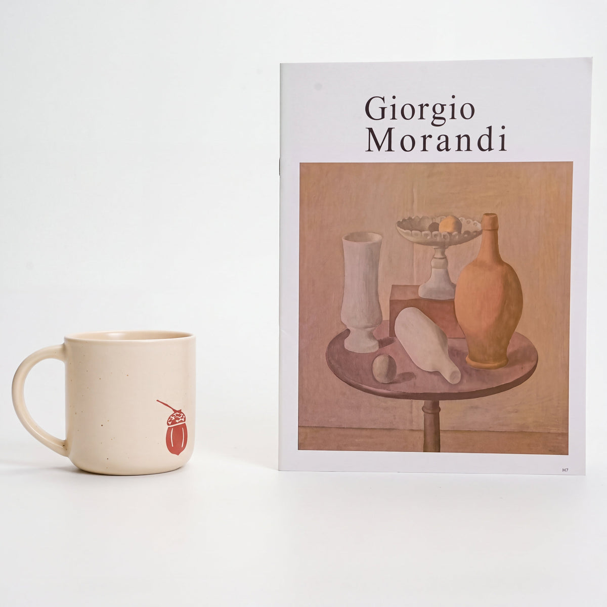 Minimalist styling of The Acorn stoneware mug showing solitary acorn motif beside Morandi art book